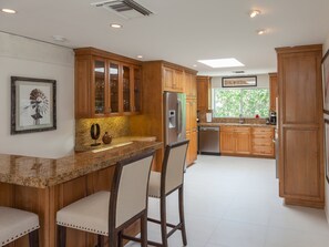 Cozinha com armários de madeira, bancada de granito, geladeira e janela com vista para árvores.