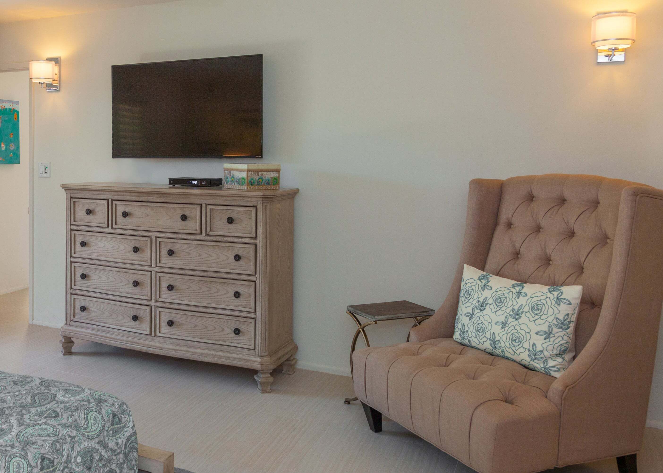 A room with a television mounted on the wall, a wooden dresser, a tufted armchair with a floral pillow, and a small table.
