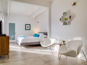 A modern hotel room with a bed, two white chairs, a small table, a wooden dresser, and a wall-mounted shelf with decorative items.