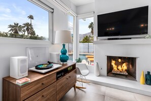 A modern living room with a fireplace, a flat-screen TV, and a view of palm trees.