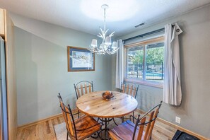 A dining area with a round wooden table, six chairs, a chandelier, a window with curtains, and a framed picture on the wall.