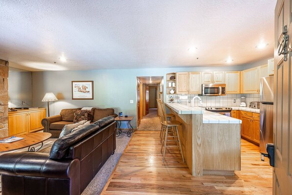A spacious living room with a leather sofa, a wooden coffee table, and a kitchen area with wooden cabinets and a stainless steel microwave.