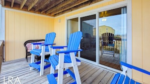 A covered outdoor area with wooden deck, blue Adirondack chairs, and a sliding glass door leading to a deck. - Dirft into Surf Condos 414 - Dolphins Drift! (Surf City)
