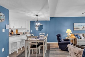 Un salon moderne avec un coin repas, agrémenté d’un mur d’accent bleu, d’un miroir rond, d’un fauteuil bleu et d’un canapé orné d’une œuvre d’art sur le thème de la plage.