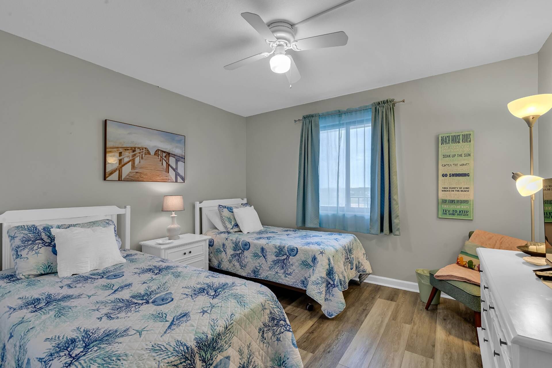 A bedroom with two beds, a ceiling fan, a painting, a window with curtains, and a sign on the wall.