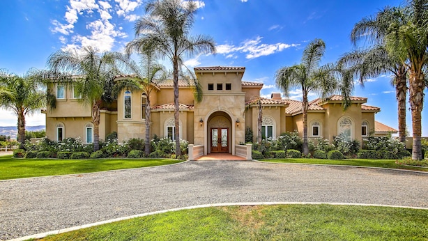 A large, two-story house with a Mediterranean-style facade, a gravel driveway, and palm trees. - Luxe Wine Country Estate w/ Orchard-Facing Balcony (Temecula)