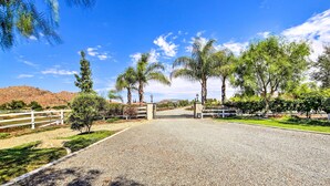A gravel driveway leading to a gated entrance with a white fence, surrounded by trees and a clear blue sky. - Luxe Wine Country Estate w/ Orchard-Facing Balcony (Temecula)