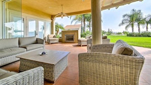 A covered outdoor seating area with wicker furniture, a fireplace, and a view of a well-maintained lawn and palm trees. - Luxe Wine Country Estate w/ Orchard-Facing Balcony (Temecula)