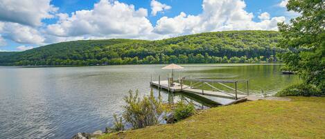 A lakeside view with a dock extending into the water, surrounded by lush greenery and a forested hill in the background.
