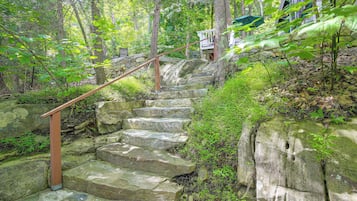 Un escalier en pierre, entouré d’une végétation luxuriante, mène à un bâtiment doté d’un mur en pierre et d’une balustrade en bois.