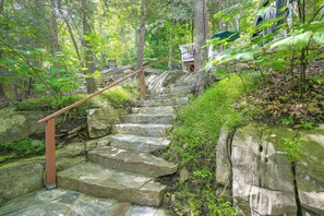 Escadaria de pedra rodeada por vegetação exuberante, levando a um edifício com parede de pedra e corrimão de madeira.