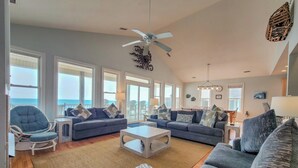 A spacious living room with a ceiling fan, large windows, and a view of the ocean.