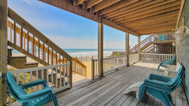 A wooden deck with a view of the beach, a staircase, and blue chairs.