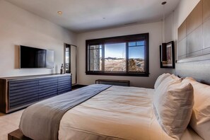 A modern bedroom with a large bed, a built-in bench, a flat-screen TV, and a window with a mountain view. - Luxurious Residence by InvitedHome | Ski-In/Ski-Out, Premier Kitchen, Modern (Edwards)