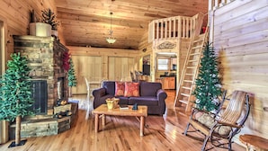 Een gezellig cabin interieur met een stenen openhaard, houten vloeren, een bank, een schommelstoel en een ladder die naar een slaapzolder leidt.