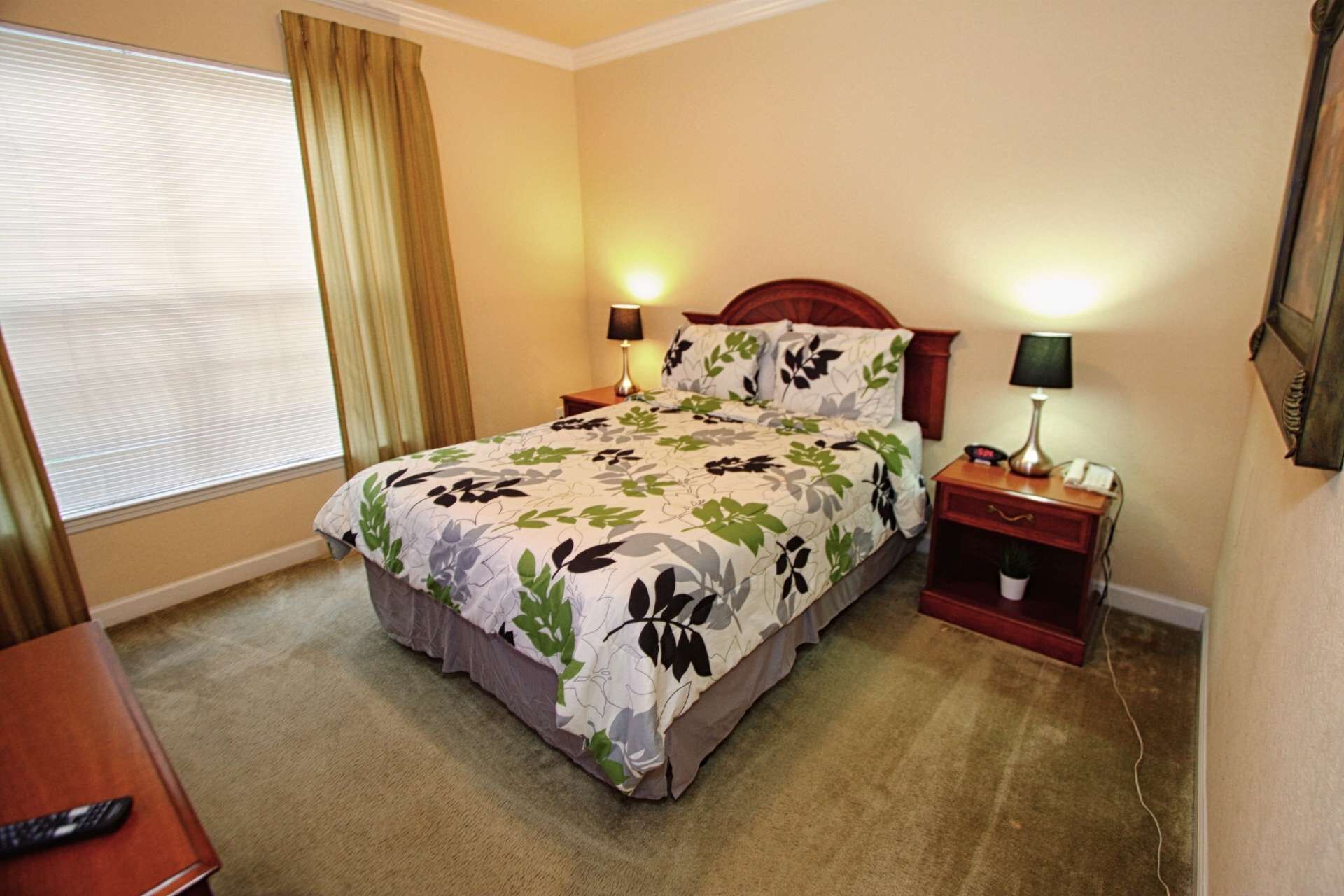 A hotel room with a bed, two bedside tables, a lamp, a nightstand, a remote control on a table, and a window with curtains.