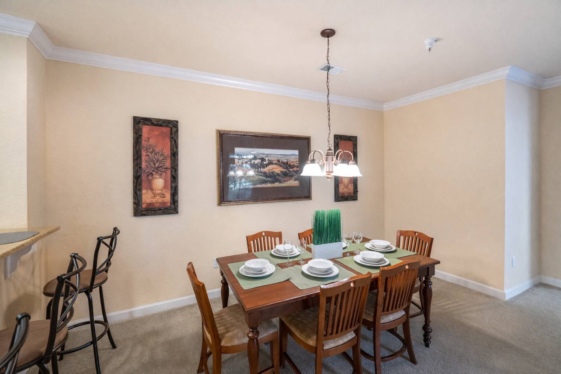 A dining area with a table set for six, surrounded by wooden chairs, a hanging light fixture, and framed artwork on the walls.