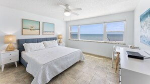 A bedroom with a bed, a desk, a chair, a ceiling fan, and a large window with a view of the ocean. - Relaxing Oceanfront Condo with Steps to Beach & Heated Pool 🌴 (Satellite Beach)