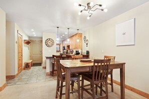 A dining area with a wooden table and chairs, a kitchen in the background, and a wall clock.