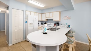 A modern kitchen with white cabinets, a central island, bar stools, and a microwave. - ETW 2005: Emerald Shore: Step Into Tranquility with a Beachfront Home! (Fort Walton Beach)