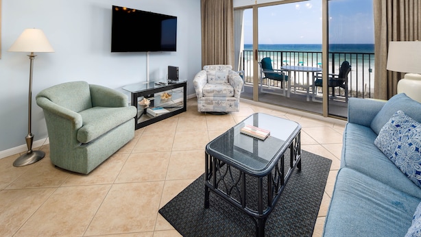 A living room with a green sofa, a blue sofa, a glass coffee table, a patterned armchair, a flat-screen TV, and a balcony with a view of the ocean. - ETW 2005: Emerald Shore: Step Into Tranquility with a Beachfront Home! (Fort Walton Beach)