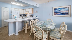 A compact kitchen with white cabinets, a refrigerator, and a microwave. There is a dining area with a glass table and wicker chairs. - ETW 2005: Emerald Shore: Step Into Tranquility with a Beachfront Home! (Fort Walton Beach)