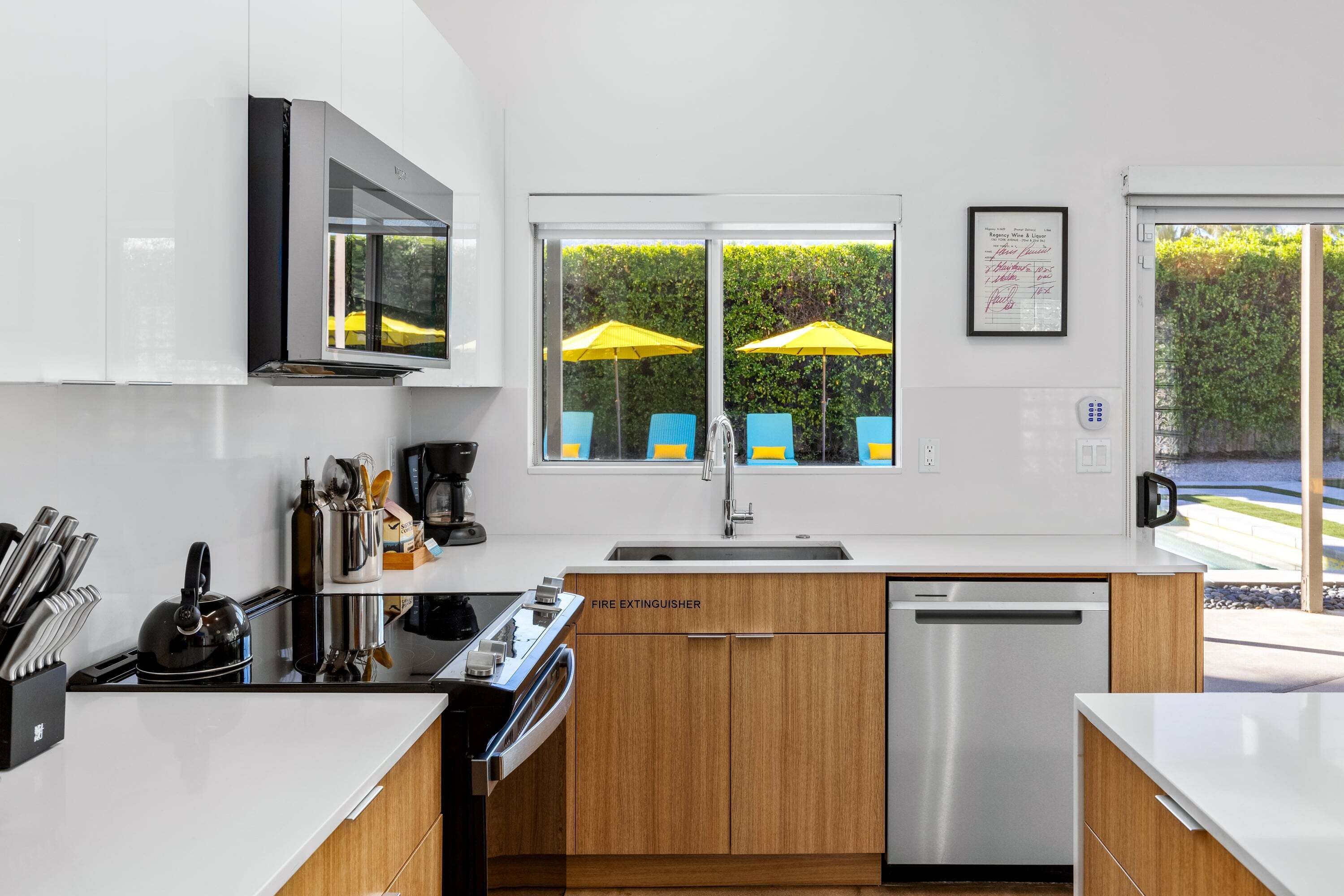 Una cocina moderna con estufa, fregadero y lavavajillas, un microondas sobre la estufa, y una ventana con paraguas amarillos afuera.