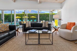 A modern living room with a black sofa, a beige armchair, and a glass coffee table. There are two yellow umbrellas outside the large windows.