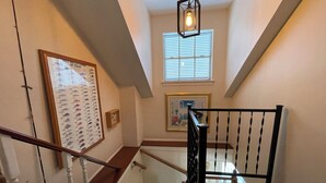 Uma escadaria com corrimão de madeira e balaústres pretos, conduzindo a um patamar com um quadro emoldurado e uma janela com persianas.