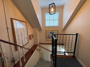 Un escalier doté d’une rampe en bois et de balustres noires, menant à un palier où se trouvent un tableau encadré et une fenêtre avec des stores.