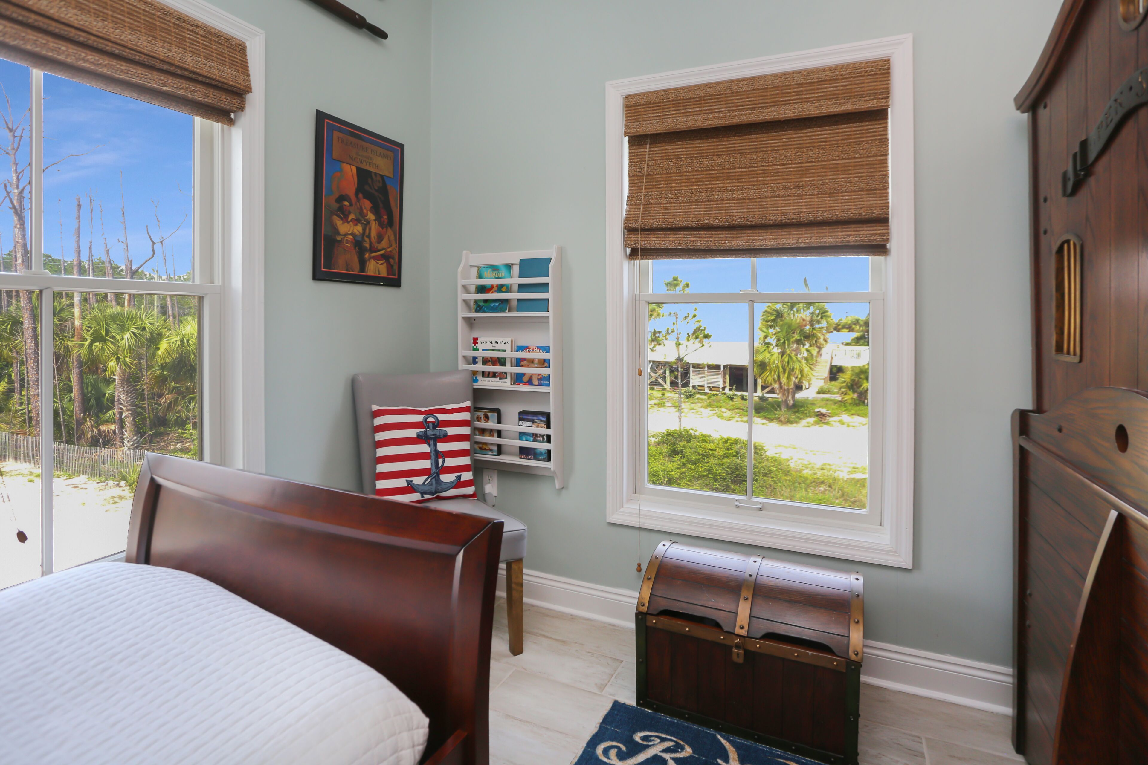 A bedroom with a large bed, a chest of drawers, a window with a view of palm trees, and a bookshelf.