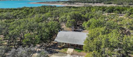 A single-story building with a metal roof, surrounded by dense greenery and a clear view of a large body of water in the background.