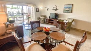 A living room with a glass table surrounded by wicker chairs, a sofa, a television, and a painting on the wall. - Hale Kamaole 340  Third floor condo, easy access to best beach on Maui! (Kihei)