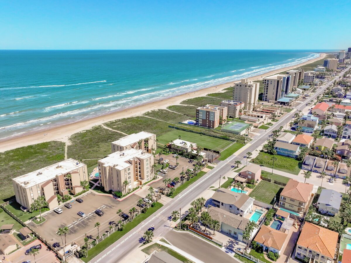 Vue aérienne d’un quartier résidentiel situé sur la côte, avec des propriétés donnant sur la plage, une route principale et un parking.