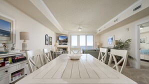 Een ruime eethoek met een witte tafel en stoelen, een plafondventilator en uitzicht op de oceaan door grote ramen.