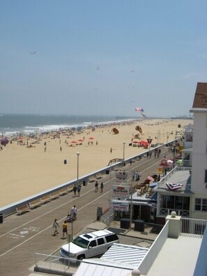 A beachside boardwalk with a white car parked on the side, a pedestrian walkway, and a view of the beach with people and umbrellas. - Enjoy a large, spacious designer unit with all the extras plus an indoor pool, outdoor pool, fitness room, assigned parking and shops on the ground level. (Ocean City)