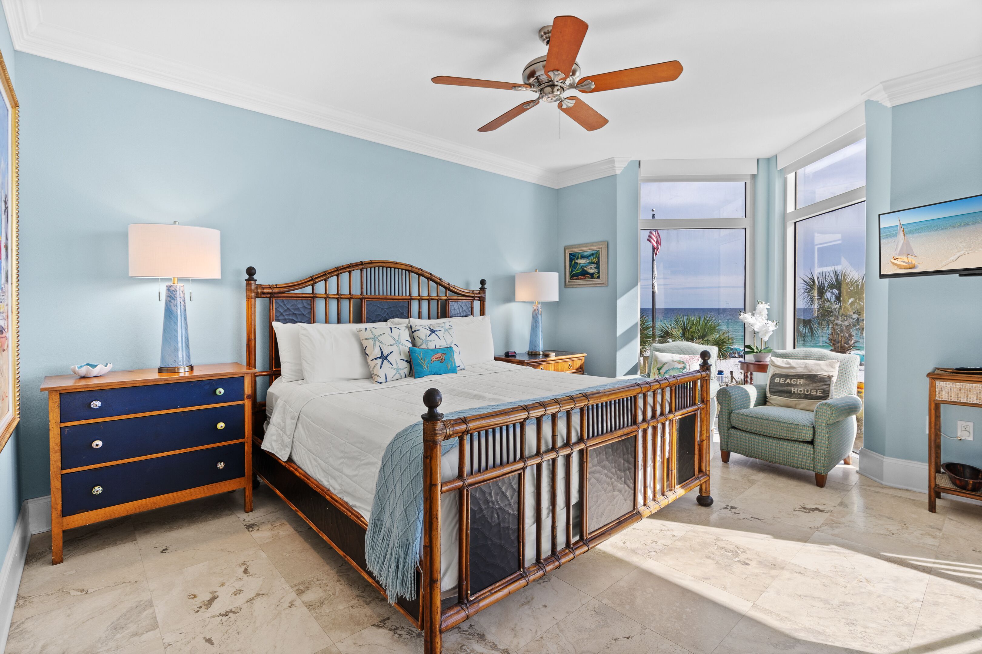 A bedroom with a wooden bed, two bedside tables, a nightstand, a chair, a ceiling fan, and a large window with a view of the ocean.