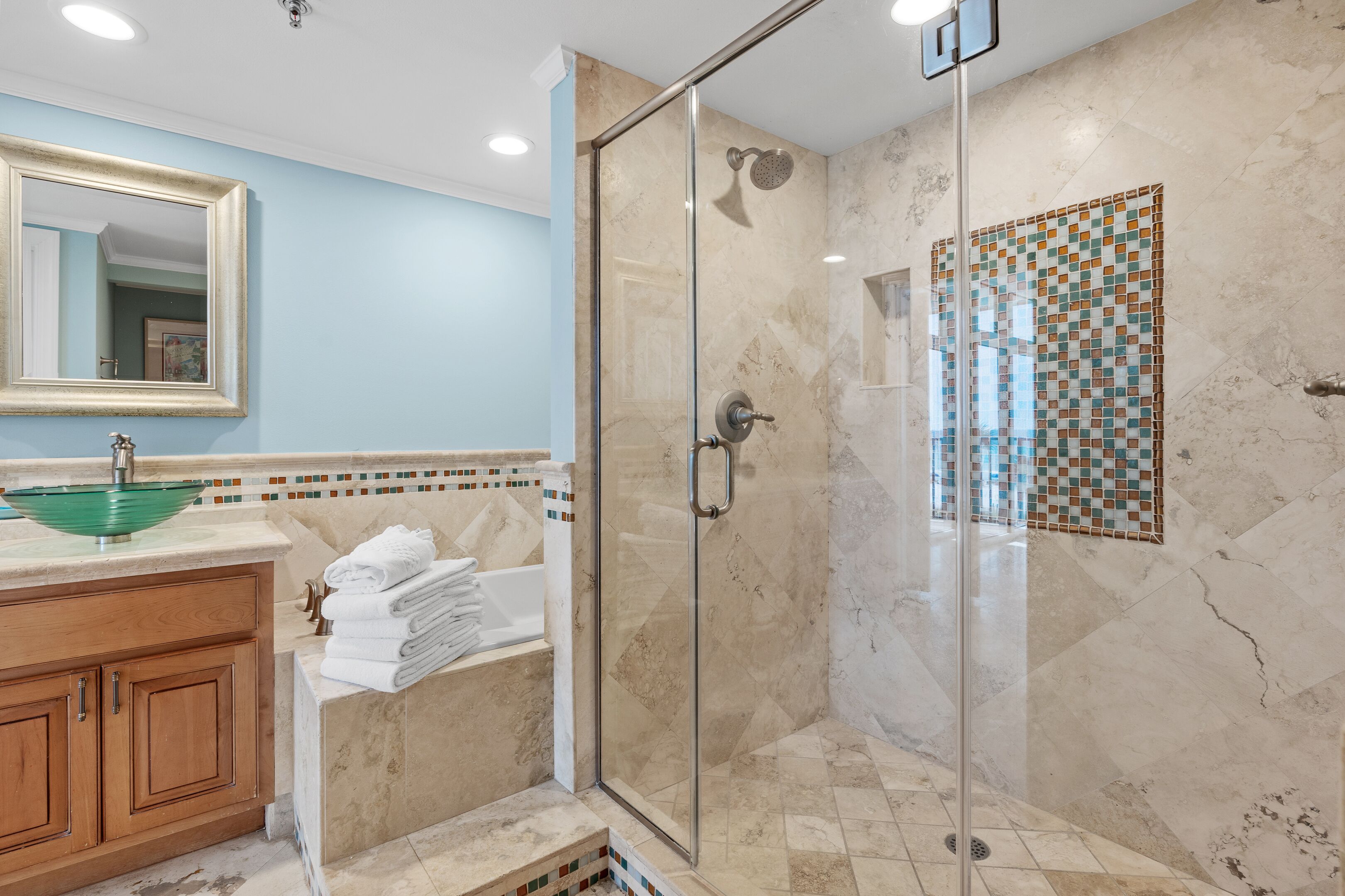 A modern bathroom with a glass-enclosed shower, a marble-tiled floor, and a wooden vanity with a green glass bowl sink.