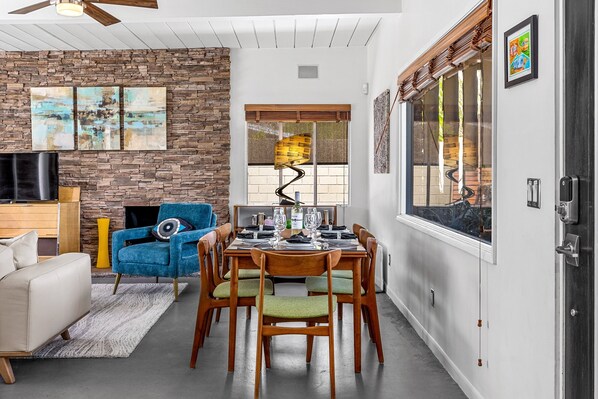 Un área de comedor con pared de piedra, un sillón azul, una mesa de madera dispuesta para cuatro personas y un televisor al fondo.