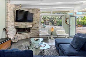 A modern living room with a stone wall, flat-screen TV, fireplace, glass coffee table, blue sofa, and a view of palm trees through large windows. - Mesa Mid-Century – Chic Poolside Escape (Palm Springs)
