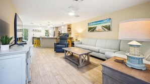 Un'ampia zona giorno con divano, tavolino e TV su un mobile. Si nota una cucina sullo sfondo con bancone e zona pranzo.