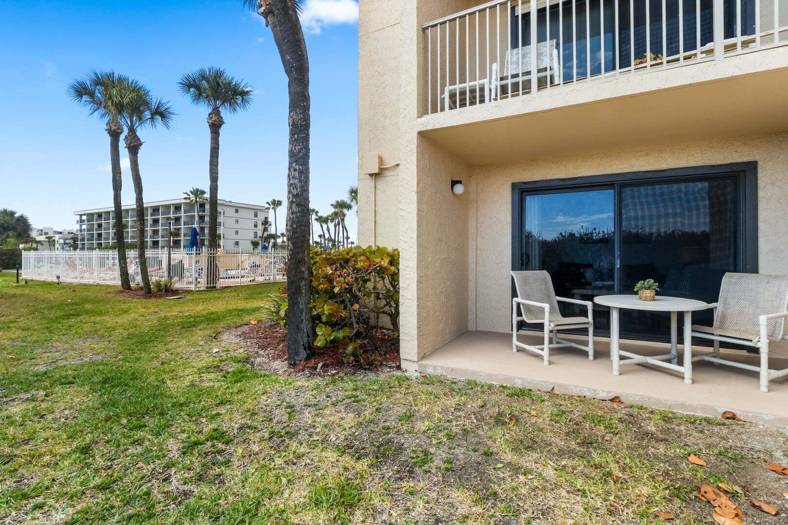 A balcony with two chairs and a small table, a palm tree, and a building in the background.