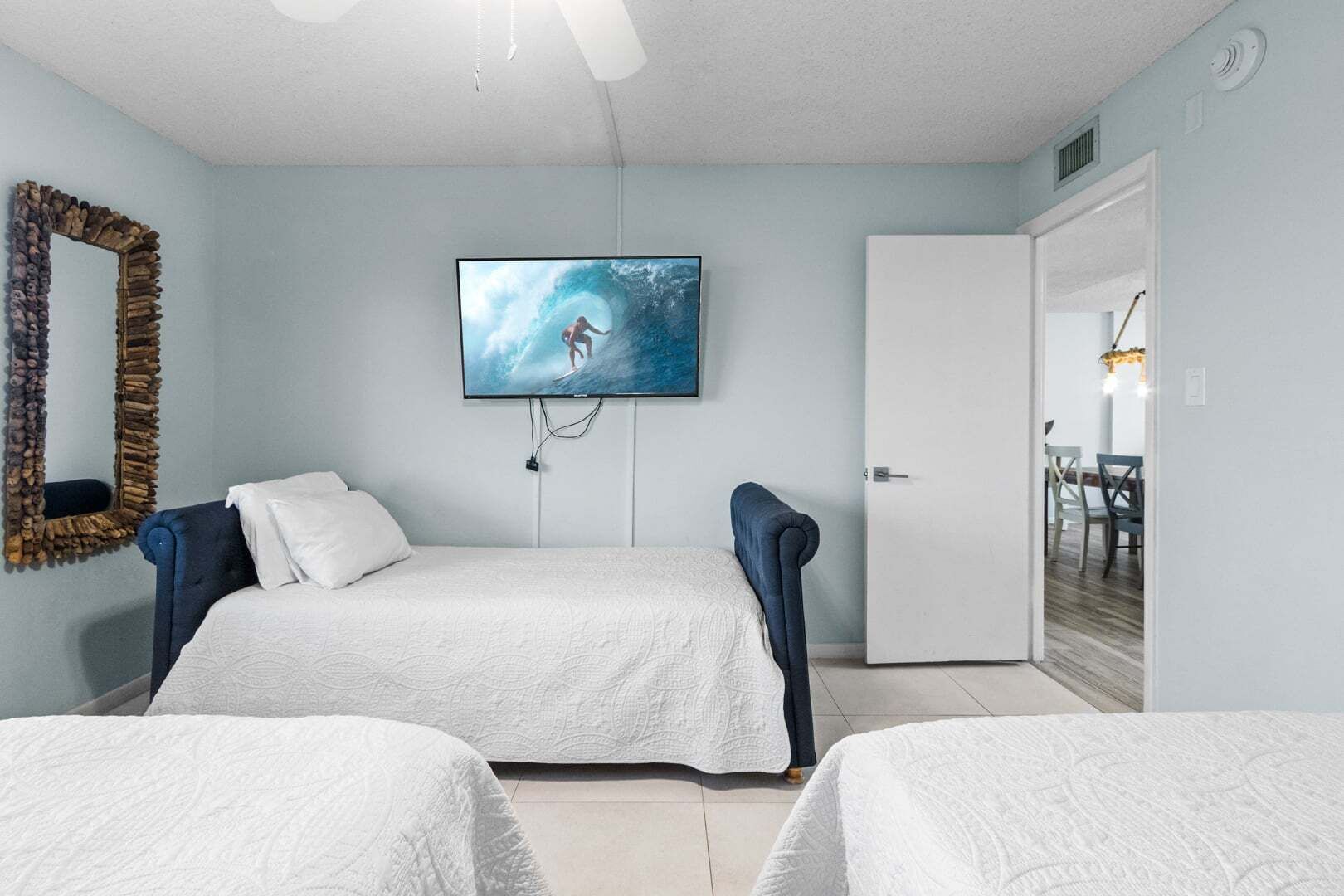 A bedroom with two beds, a TV on the wall, a mirror, and a door leading to a dining area.