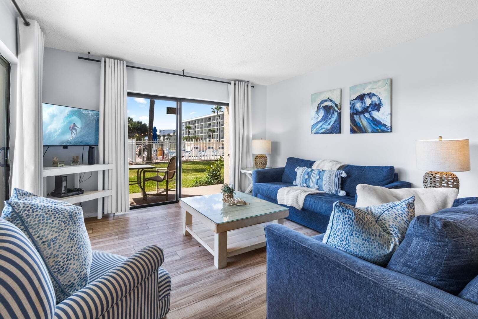 A living room with a TV, blue sofas, a coffee table, and a view of a building and greenery.
