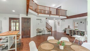 A spacious dining area with a wooden table and white chairs, a staircase leading to an upper level, and a kitchen area in the background. - Family Home w/ Deck ~ 2 Mi to Camelback Mountain! (Tannersville)