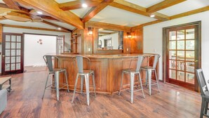 A spacious room with a wooden bar and four bar stools. The room has wooden beams on the ceiling and a large window with a view of the outdoors. - Family Home w/ Deck ~ 2 Mi to Camelback Mountain! (Tannersville)