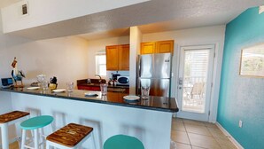 A kitchen with a central island, wooden stools, a refrigerator, and a door leading to a patio. - Beachfront on Pass-A-Grille Beach with Pool! Walking Distance to Great Restaurants and Shops. (St. Pete Beach)