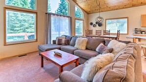 Una sala de estar acogedora con un sofá modular, una mesa de centro de madera y grandes ventanales con vistas a árboles.