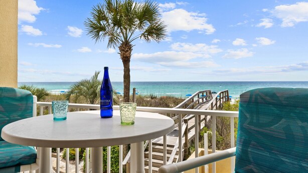 A balcony with a table, chairs, a palm tree, and a view of the ocean. - SL 101: Sugary Sands - Cute and Cozy with Beautiful Upgrades!! (Fort Walton Beach)