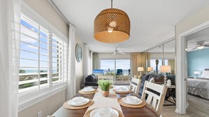 A dining area with a table set for four, a hanging woven light fixture, and a view of the ocean through large windows. - SL 101: Sugary Sands - Cute and Cozy with Beautiful Upgrades!! (Fort Walton Beach)